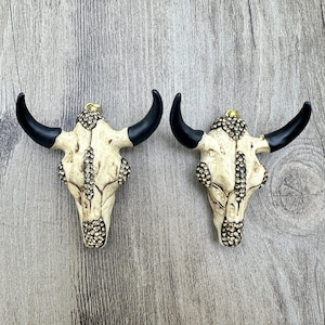May include: Two decorative bull skull pendants. The skulls are off-white with black horns and are embellished with sparkling rhinestones. The pendants have a gold-coloured loop at the top for hanging. The background is a light grey wood grain.