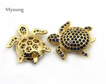 May include: Two gold-tone turtle charms with blue crystal accents. The turtle on the left has a honeycomb design on its shell. The turtle on the right has a solid shell with blue crystals.