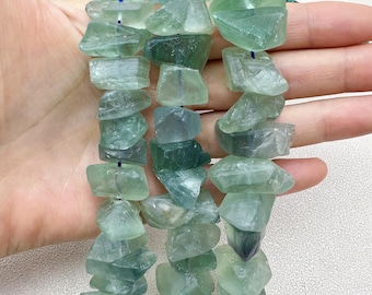 May include: Strands of raw, uncut fluorite gemstone beads in shades of green and blue-green. The beads are irregular in shape and strung together with a dark blue string. The gemstones are translucent and have a natural, rough texture.