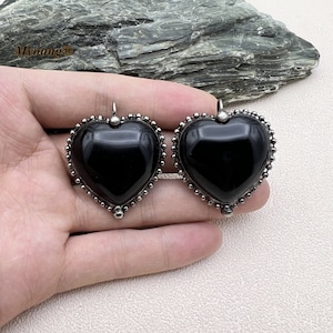 Puede incluir: Un par de pendientes de corazón negro con engastes de metal plateado. Los pendientes tienen un borde de cuentas y un acabado liso y pulido.