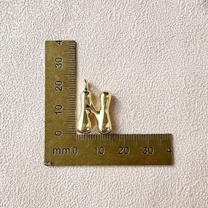 May include: A gold-colored pendant in the shape of the letter "N" is displayed next to a ruler. The pendant is approximately 1 inch in height. The ruler shows measurements in millimeters.