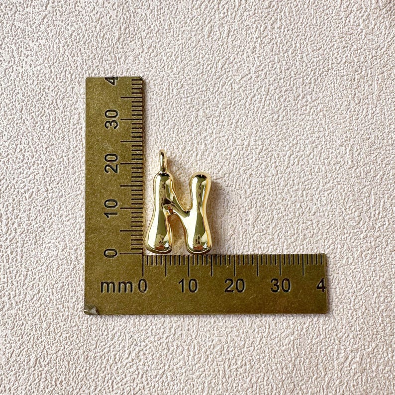 May include: A gold-colored pendant in the shape of the letter "N" is displayed next to a ruler. The pendant is approximately 1 inch in height. The ruler shows measurements in millimeters.