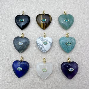 May include: Nine heart-shaped pendants in various colors, including black, brown, green, gray, white, blue, and purple. Each pendant features a gold-colored bail and a small, oval-shaped gemstone in the center. The pendants are displayed on a light-colored surface.