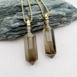 Pode incluir: Dois colares de corrente dourada com pingentes de garrafas de cristal de quartzo fumado. As garrafas têm uma tampa dourada e um inserto de vidro transparente.
