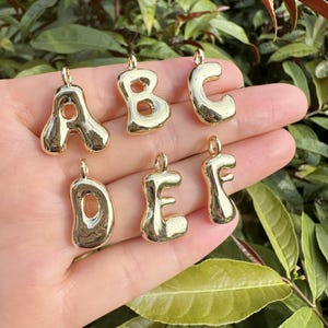 May include: Gold-colored alphabet charms, including the letters A, B, C, D, E, and F. Each charm has a rounded, bubbly design with a small loop at the top for attaching to a necklace or bracelet. The charms are displayed against a green background.