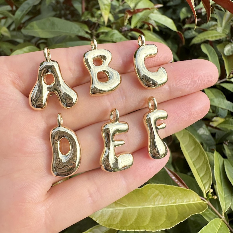 May include: Gold-colored alphabet charms, including the letters A, B, C, D, E, and F. Each charm has a rounded, bubbly design with a small loop at the top for attaching to a necklace or bracelet. The charms are displayed against a green background.