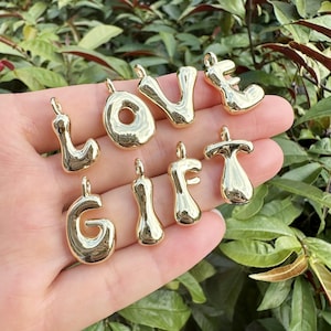May include: Gold-colored metal charms spelling out the words "LOVE" and "GIFT". The charms are in a bubbly, rounded font, each with a small loop at the top for attaching to a chain or bracelet. The charms are displayed against a background of green foliage.