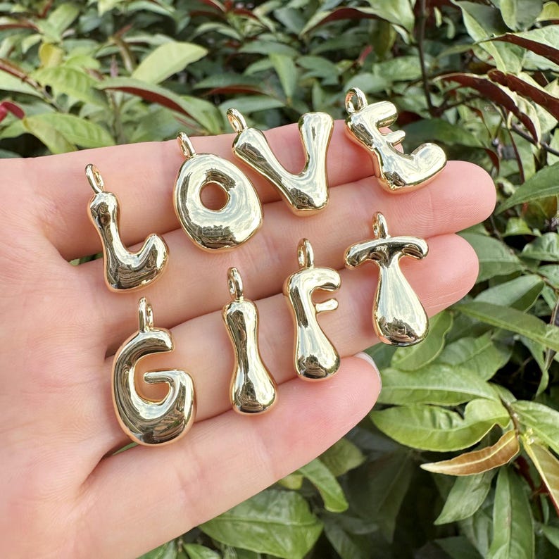 May include: Gold-colored metal charms spelling out the words "LOVE" and "GIFT". The charms are in a bubbly, rounded font, each with a small loop at the top for attaching to a chain or bracelet. The charms are displayed against a background of green foliage.