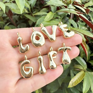May include: Gold-colored metal charms spelling out "LOVE" and "GIFT". The charms are in a bubbly, rounded font, each with a small loop at the top for attaching to a chain or bracelet. The background is a blur of green foliage.