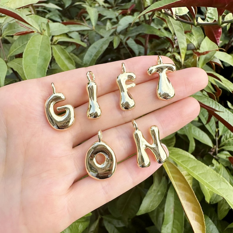 May include: Gold-colored alphabet charms, including the letters G, I, F, T, O, and H. The charms are shiny and have a rounded, bubbly design. They are displayed against a background of green foliage.