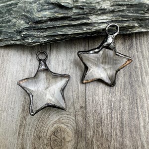 May include: Two silver-toned metal star-shaped pendants with clear glass inserts. The pendants are hanging on a wooden surface.