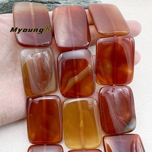 May include: A strand of carnelian beads, each bead is a rectangular shape with a smooth, polished finish. The beads are a mix of orange, red, and brown colours with natural agate banding.