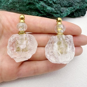 Può includere: Due flaconi di profumo in cristallo grezzo trasparente con tappi color oro. I flaconi hanno una consistenza ruvida e naturale. I tappi hanno un piccolo design arrotondato. I flaconi sono esposti su uno sfondo chiaro.