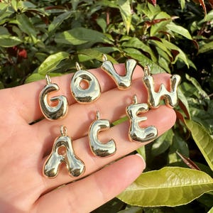 May include: Gold-colored alphabet charms, including the letters G, O, V, W, A, C, and E. Each charm has a rounded, bubble-like design with a small loop at the top for attaching to a chain or bracelet. The charms are displayed against a background of green foliage.