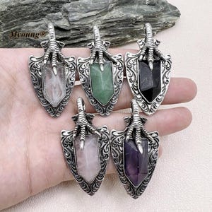 Puede incluir: Cinco colgantes de flecha de plata con piedras preciosas de diferentes colores. Los colgantes presentan un diseño detallado con una garra en la parte superior. Las piedras preciosas son transparentes, verdes, negras, rosas y moradas.