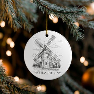 May include: A white ceramic Christmas ornament with a black and white illustration of a windmill. The text "EAST HAMPTON, NY" is printed below the windmill. The ornament hangs from a gold string.