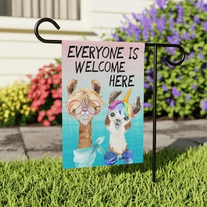 May include: A garden flag featuring the phrase "EVERYONE IS WELCOME HERE" and two llama illustrations. One llama wears glasses and a scarf, the other a unicorn horn and bow tie. The background is a pink and blue gradient.