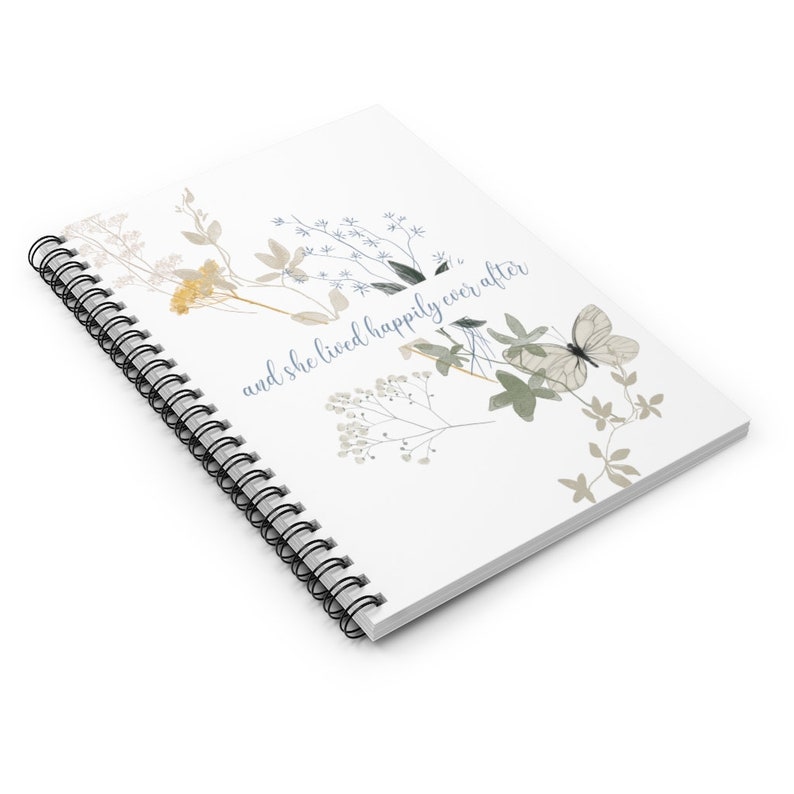 K&ouml;nnte beinhalten: Ein wei&szlig;es Spiralnotizbuch mit einem floralen Design und dem Text "and she lived happily ever after". Die Blumen sind in Blaut&ouml;nen, Gr&uuml;n- und Braunt&ouml;nen gehalten.