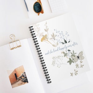 K&ouml;nnte beinhalten: Ein wei&szlig;es Notizbuch mit Spiralbindung, einem floralen Design und dem Text "and she lived happily ever after".