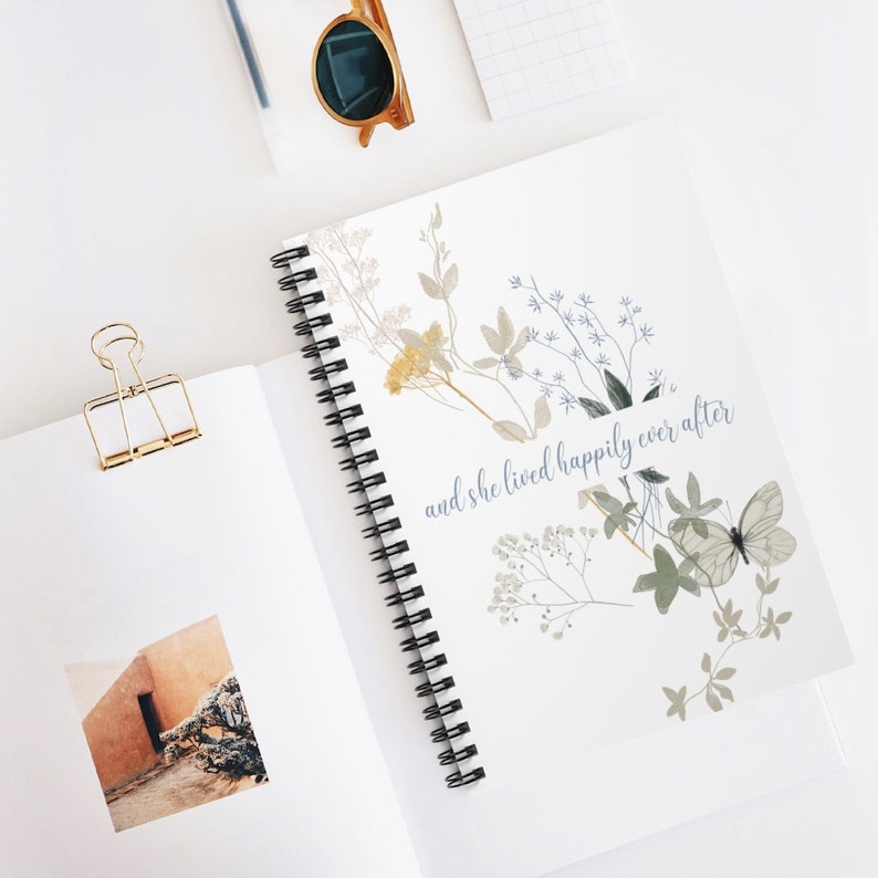 K&ouml;nnte beinhalten: Ein wei&szlig;es Notizbuch mit Spiralbindung, einem floralen Design und dem Text "and she lived happily ever after".