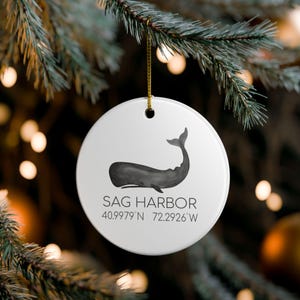 Può includere: Ornamento natalizio rotondo in ceramica bianca con un'illustrazione di balena nera. L'ornamento presenta il testo "SAG HARBOR" e le coordinate 40.9979°N 72.2926°W. È appeso a un filo dorato. L'ornamento è su un albero di Natale.