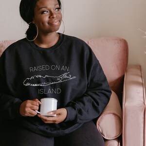 May include: A person wearing a black sweatshirt with the text "RAISED ON AN ISLAND" and a white outline of Long Island. The person is holding a white enamel mug.