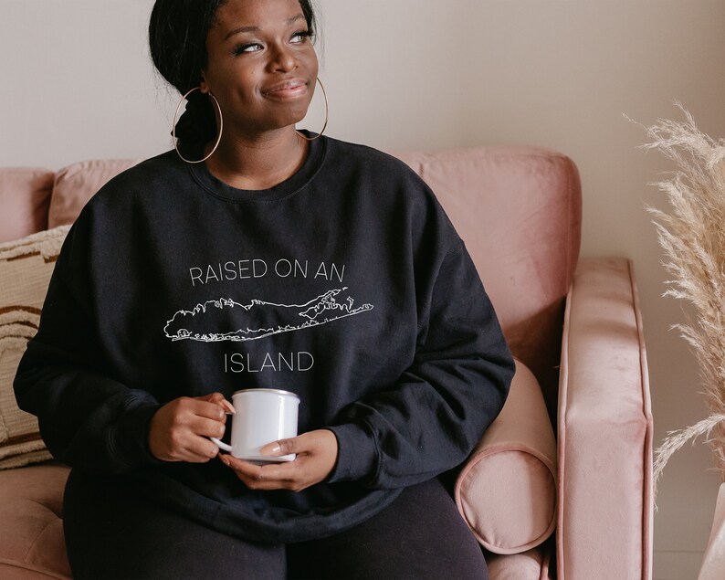 May include: A person wearing a black sweatshirt with the text "RAISED ON AN ISLAND" and a white outline of Long Island. The person is holding a white enamel mug.