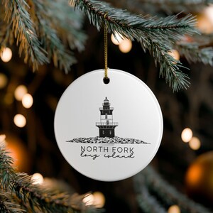 May include: A white ceramic Christmas ornament with a black and white illustration of a lighthouse on a rocky base. The text "North Fork Long Island" is below the lighthouse. The ornament hangs from a gold string.