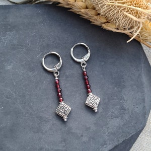 May include: A pair of silver-toned hoop earrings featuring a vertical drop design. Each earring consists of a small silver hoop attached to a string of faceted deep red garnet beads, ending in a square silver-toned charm with a floral pattern.
