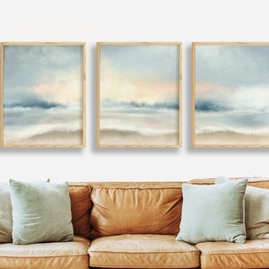 Pu&ograve; includere: Tre stampe artistiche di paesaggi incorniciate. Ogni stampa presenta una rappresentazione morbida e astratta di una scena costiera con sfumature blu, beige e rosa tenui. L'opera d'arte &egrave; esposta sopra un divano in pelle marrone con cuscini azzurri.