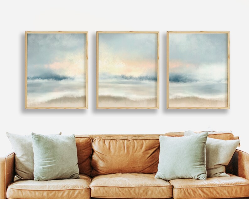 Pu&ograve; includere: Tre stampe artistiche di paesaggi incorniciate. Ogni stampa presenta una rappresentazione morbida e astratta di una scena costiera con sfumature blu, beige e rosa tenui. L'opera d'arte &egrave; esposta sopra un divano in pelle marrone con cuscini azzurri.