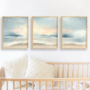 Pu&ograve; includere: Tre stampe artistiche incorniciate di paesaggi. Ogni stampa presenta una serena scena di spiaggia con un cielo azzurro chiaro, un tocco di pesca e una riva sabbiosa. L'opera d'arte &egrave; esposta sopra due cuscini decorativi color crema e una culla di legno.