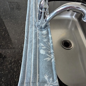 May include: A light blue kitchen towel with white floral and striped patterns is draped over a chrome faucet. The towel is made of a soft, absorbent material and is designed to catch water drips. The sink is stainless steel.