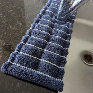 May include: A navy blue absorbent kitchen sink protector with white horizontal stripes. The rectangular pad is designed to fit around a faucet and protect the sink area from water spots and scratches.