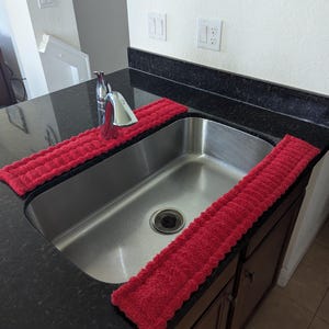 May include: A stainless steel kitchen sink with a chrome faucet and two red absorbent mats. The mats are placed on the black countertop on either side of the sink. The sink is set against a white wall.