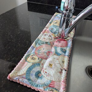 May include: A kitchen sink with a chrome faucet and a decorative dish towel with a pink, blue, and gold umbrella pattern. The towel has a pink crocheted border.