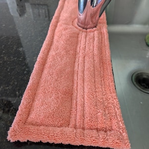 May include: A coral-colored microfiber sink mat with a hole for a faucet. The mat is placed on a black countertop.