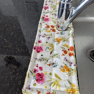 May include: A white absorbent kitchen towel with a floral pattern of pink, red, orange, and purple flowers. The towel is placed around a chrome faucet, with a yellow trim.