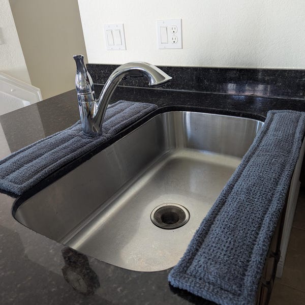 May include: A stainless steel kitchen sink with a chrome faucet and two dark gray, rectangular, textured absorbent fabric mats placed along the edges of the black granite countertop to catch water drips around the basin area.