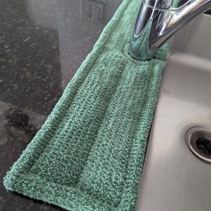 May include: A sage green rectangular absorbent fabric mat designed to fit around the base of a kitchen faucet. The mat features a textured, terry-cloth style surface with stitched edges and a central cutout to accommodate the faucet neck.