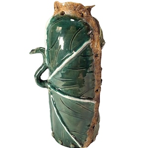 May include: A green ceramic vase with a leaf design and a brown textured rim. The vase has a unique shape with a handle that resembles a leaf.