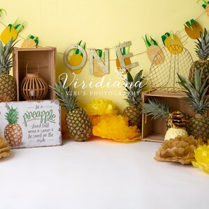 Può includere: Uno sfondo per feste di primo compleanno a tema ananas giallo e bianco con una cassa di legno, un cartello a forma di ananas che dice "be a pineapple stand tall wear a crown & be sweet on the inside", un uno dorato e fiori di carta velina gialli e marroni.