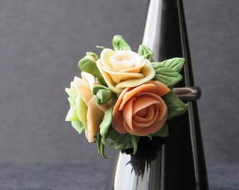 May include: A ring with a cluster of three peach and cream colored fabric roses with green leaves. The ring is displayed on a black ring holder.