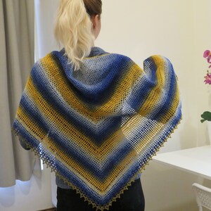 May include: A handmade, open-weave shawl in shades of blue, yellow, and gray. The shawl features a striped pattern and a scalloped edge. The shawl is draped over the shoulders.