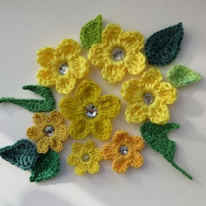 May include: A collection of crocheted yellow flowers with green leaves and sparkly centers. The flowers are arranged in a circular pattern on a white background.