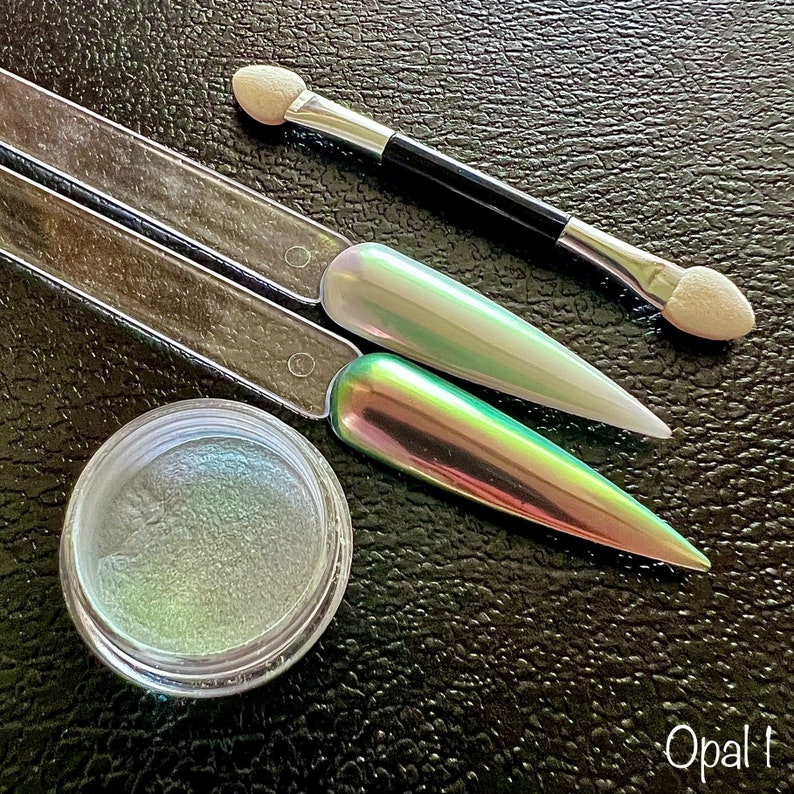 May include: A small jar of iridescent green and white nail polish pigment with two applicators and the word "Opal" written on the bottom of the image.
