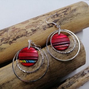 Puede incluir: Un par de pendientes de aro de plata con un colgante circular de rayas rojas y negras. Los pendientes tienen un diseño de aro doble.