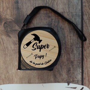 Può includere: Un metro a nastro nero e marrone con un viso in legno. Il viso ha un design a martello e chiodo nero e il testo "Super Pappy! de la part de Gabin".