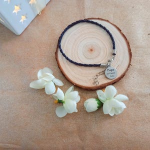 Peut inclure: Un bracelet en cuir noir avec un charme argenté qui dit "Papa je t'aime" sur une surface en bois avec des fleurs blanches.