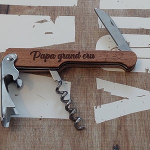 May include: A wooden multi-tool with a corkscrew, bottle opener, and a small knife. The handle is engraved with the words "Papa grand cru" and two small hearts.